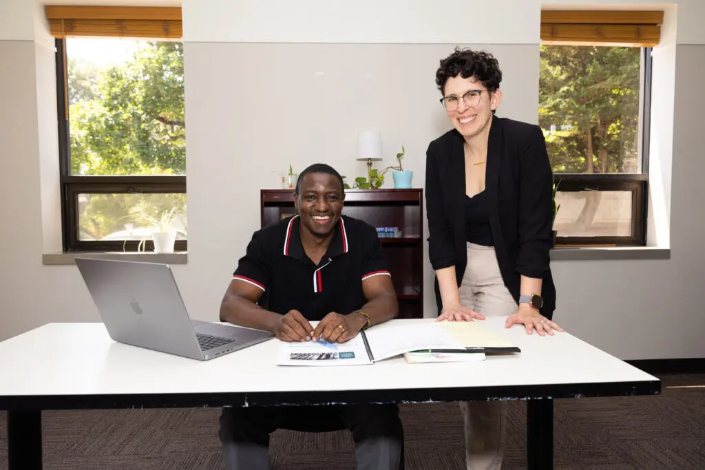 A man seated at a desk in a classroom with a laptop and books. A female teacher stands next to him.