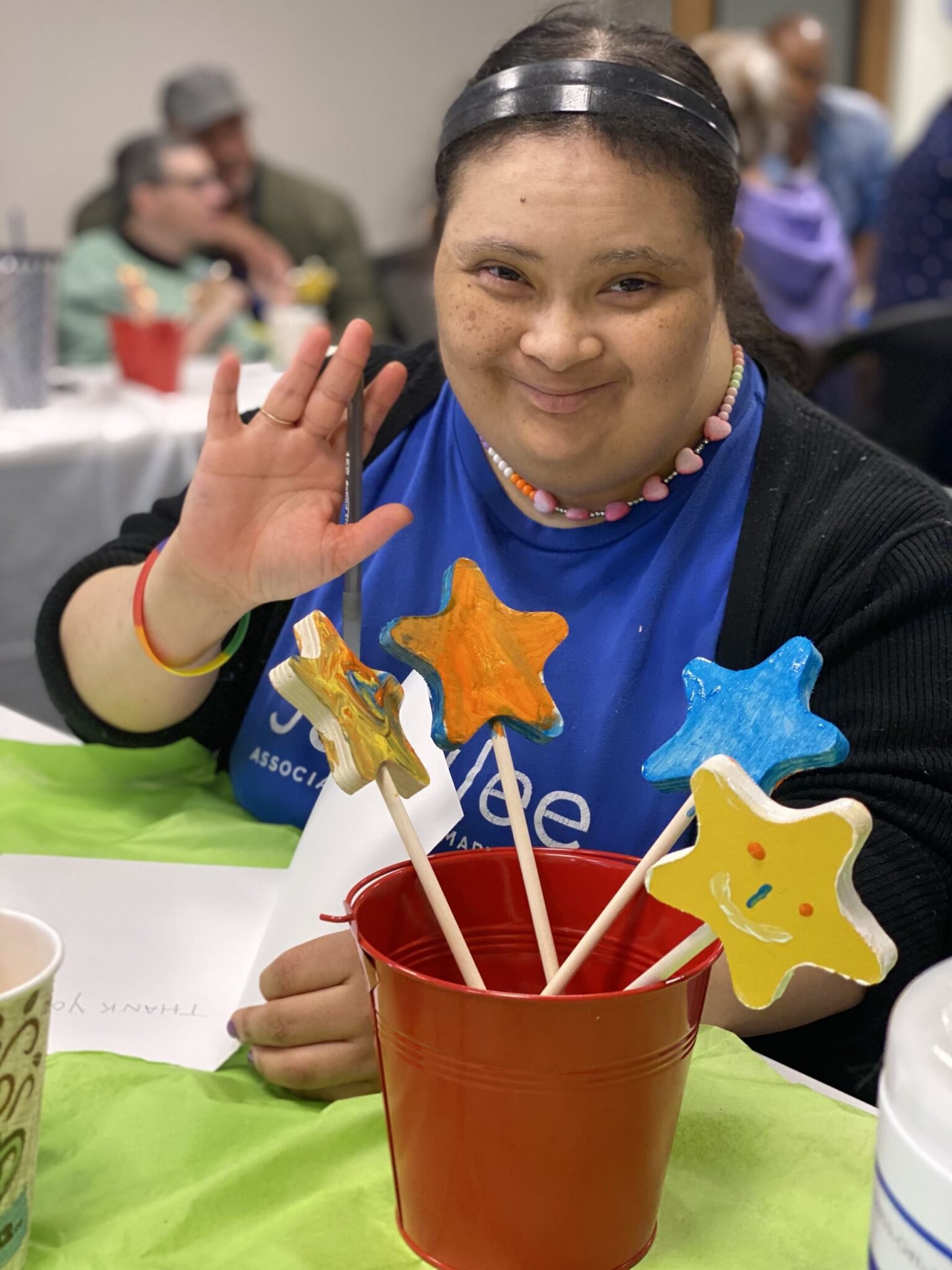 A woman smiles and waves with a paintbrush in her hand