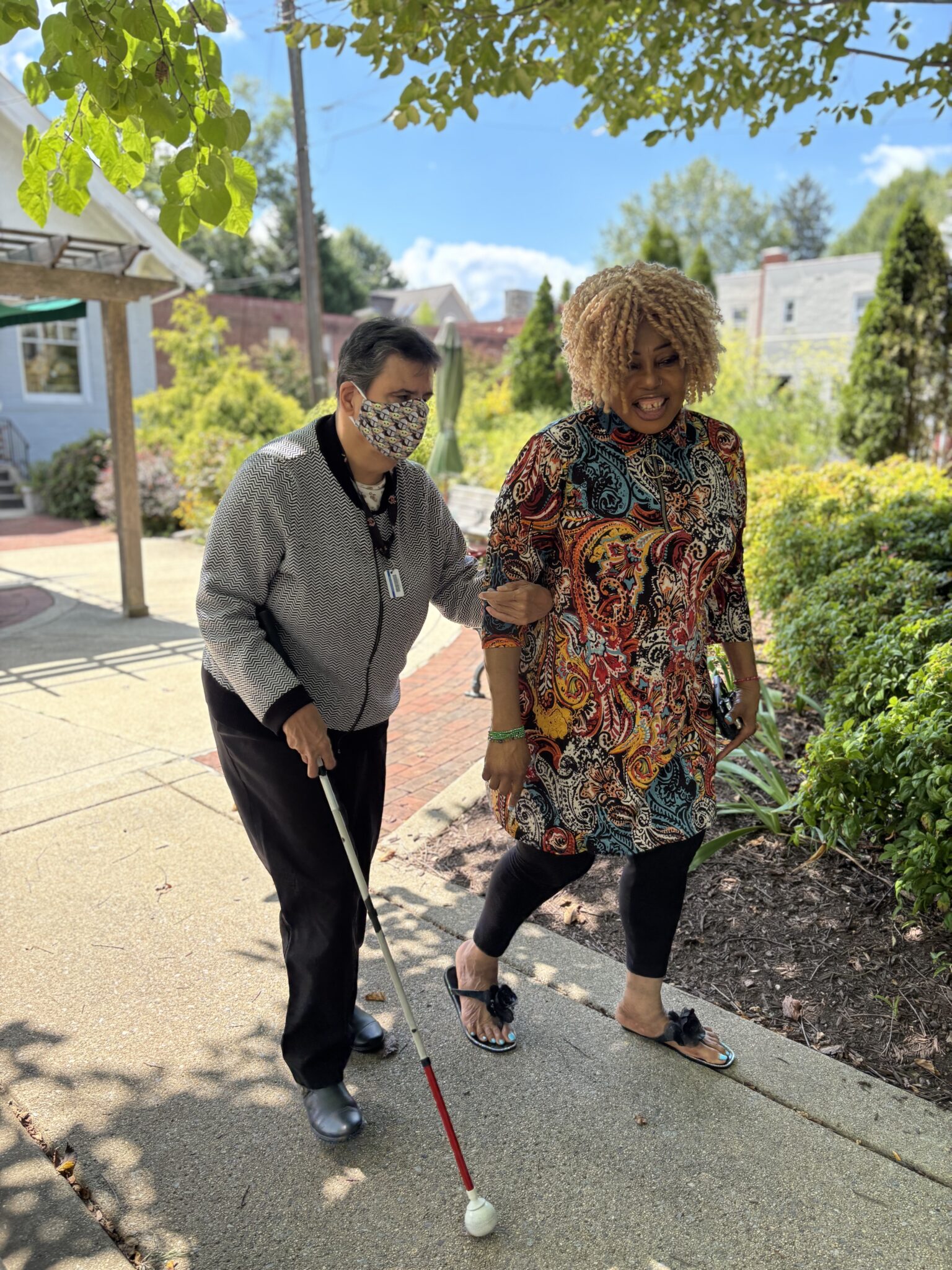 Two women, one with a cane, walk arm in arm outside on a sunny day.