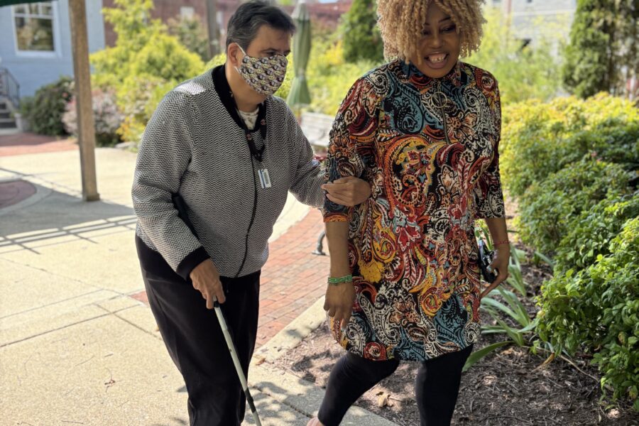 Two women, one with a cane, walk arm in arm outside on a sunny day.