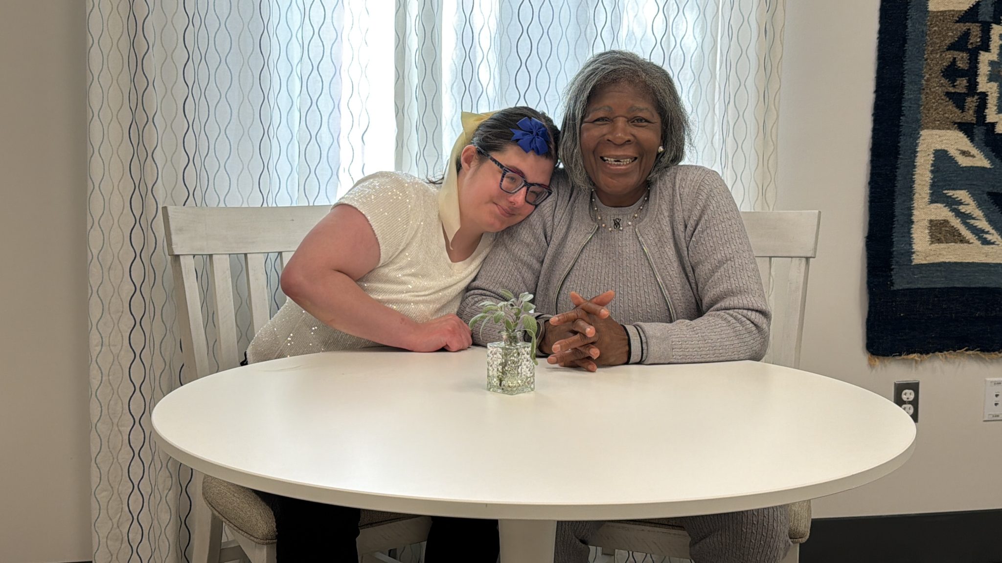 Two women sit at a table together. The younger woman affectionately rests her head on the other woman's shoulder.