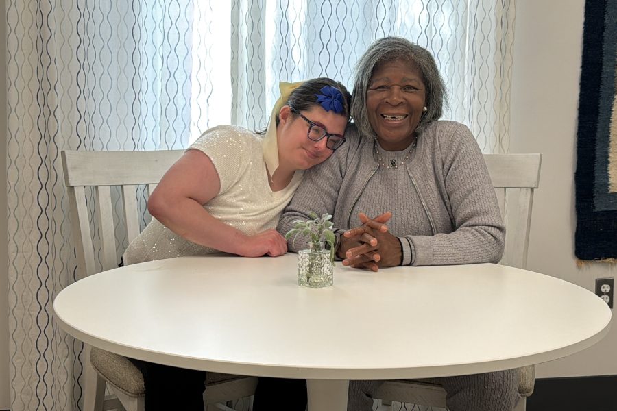 Two women sit at a table together. The younger woman affectionately rests her head on the other woman's shoulder.