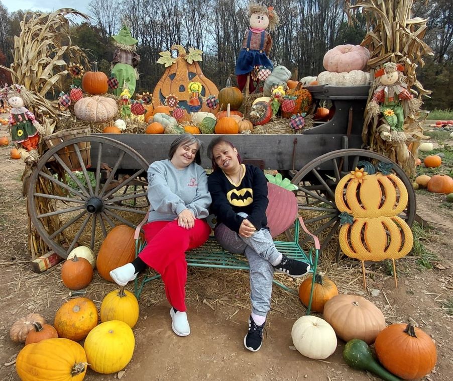 Two women sit together smiling in the middle of a pumpkin patch