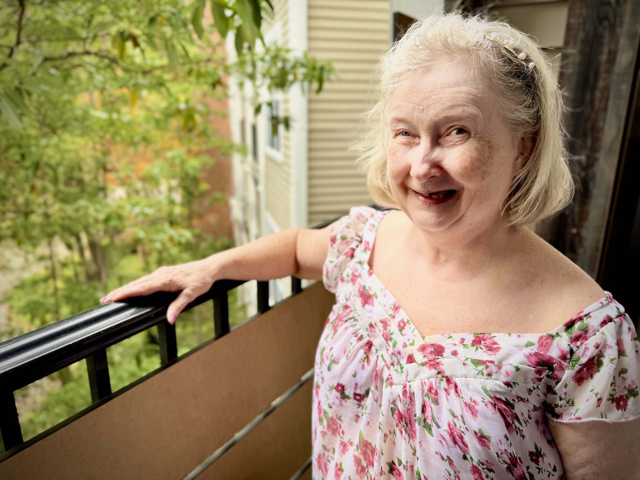 An older woman stands on her apartment balcony and smiles