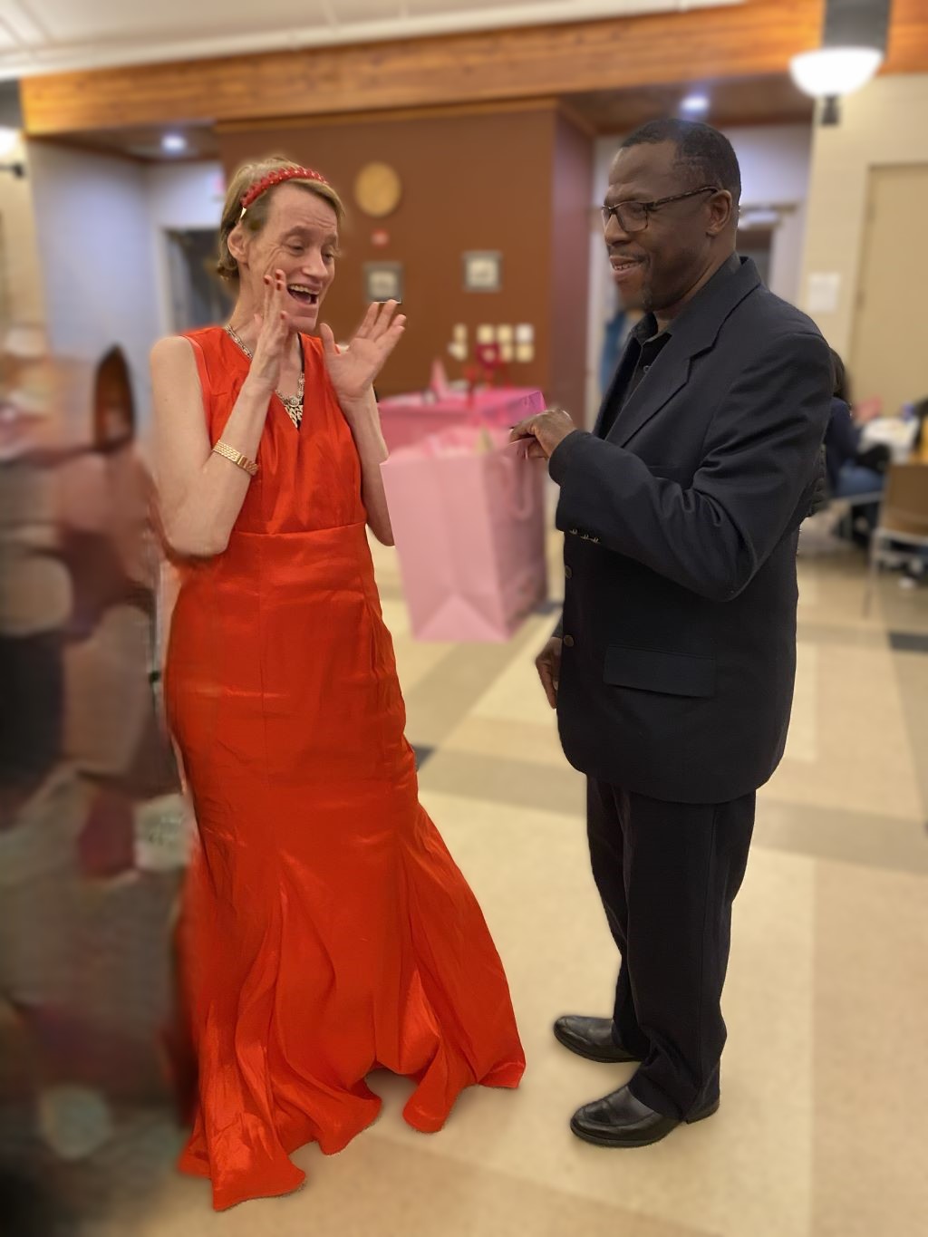 A lady in a red dress is surprised by being gifted a Valentine's Day present at a party.